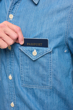 Hand placing a passport into the chech pocket of a blue chambray shirt.